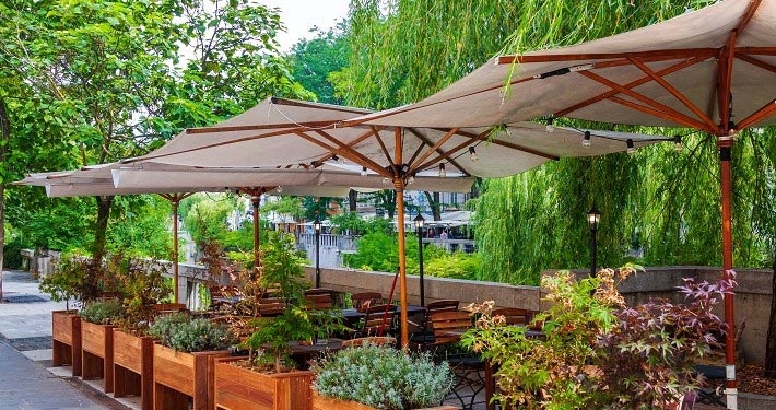 Weißer Sonnenschirm für die Gastro – auf einer Terrasse vor Bäumen an einem Fluss.